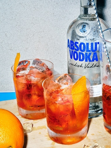 Imagem de dois copos de drinks com vodka, gelo e twist de laranja, ao lado de uma jarra com os mesmos itens e mais uma colher bailarina, uma garrafa de vodka Absolut e uma laranja na frente.