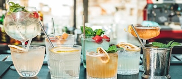 Imagem de uma mesa com dois tipos de taças contendo drinks de diferentes tamanhos e sabores, além de outros copos de drinks com canudos de metal e diferentes sabores.
