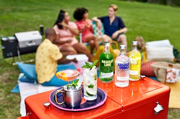 Imagem de três garrafas de vodka Absolut, uma original e as outras saborizadas, ao lado de um prato com drinks, sobre uma caixa térmica vermelha. Ao fundo, pessoas sentadas conversando ao ar livre.